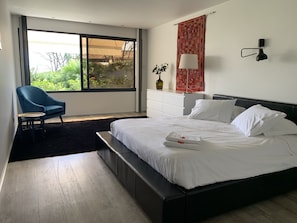 Ein modernes Hotelzimmer mit einem großen Bett, einem blauen Sessel, einem kleinen Tisch, einer Kommode und einem Fenster mit Blick auf Grünflächen.
