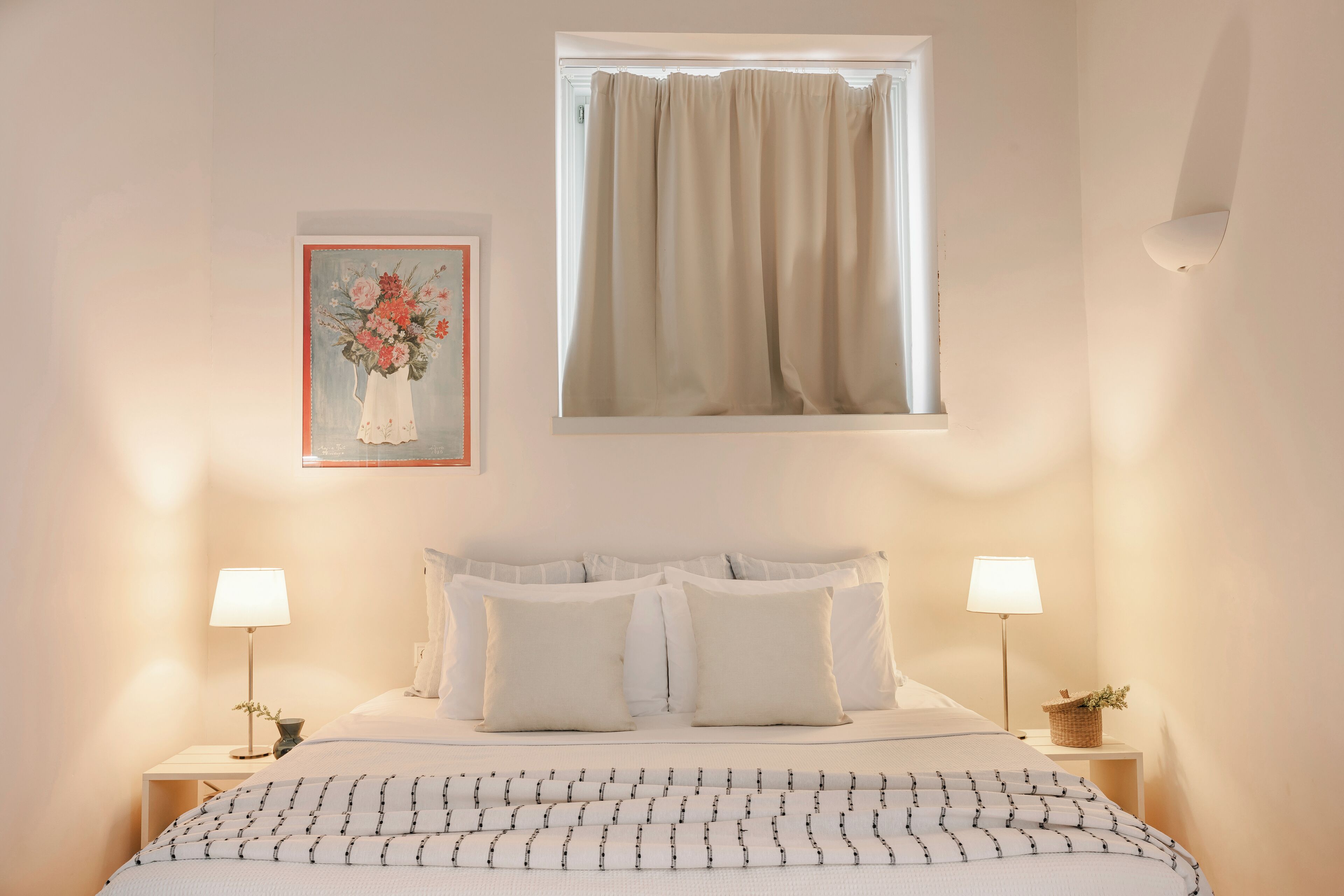 A neatly arranged bedroom with a bed, two bedside tables with lamps, a framed floral artwork, and a window with curtains.