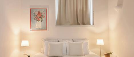 A neatly arranged bedroom with a bed, two bedside tables with lamps, a framed floral artwork, and a window with curtains.