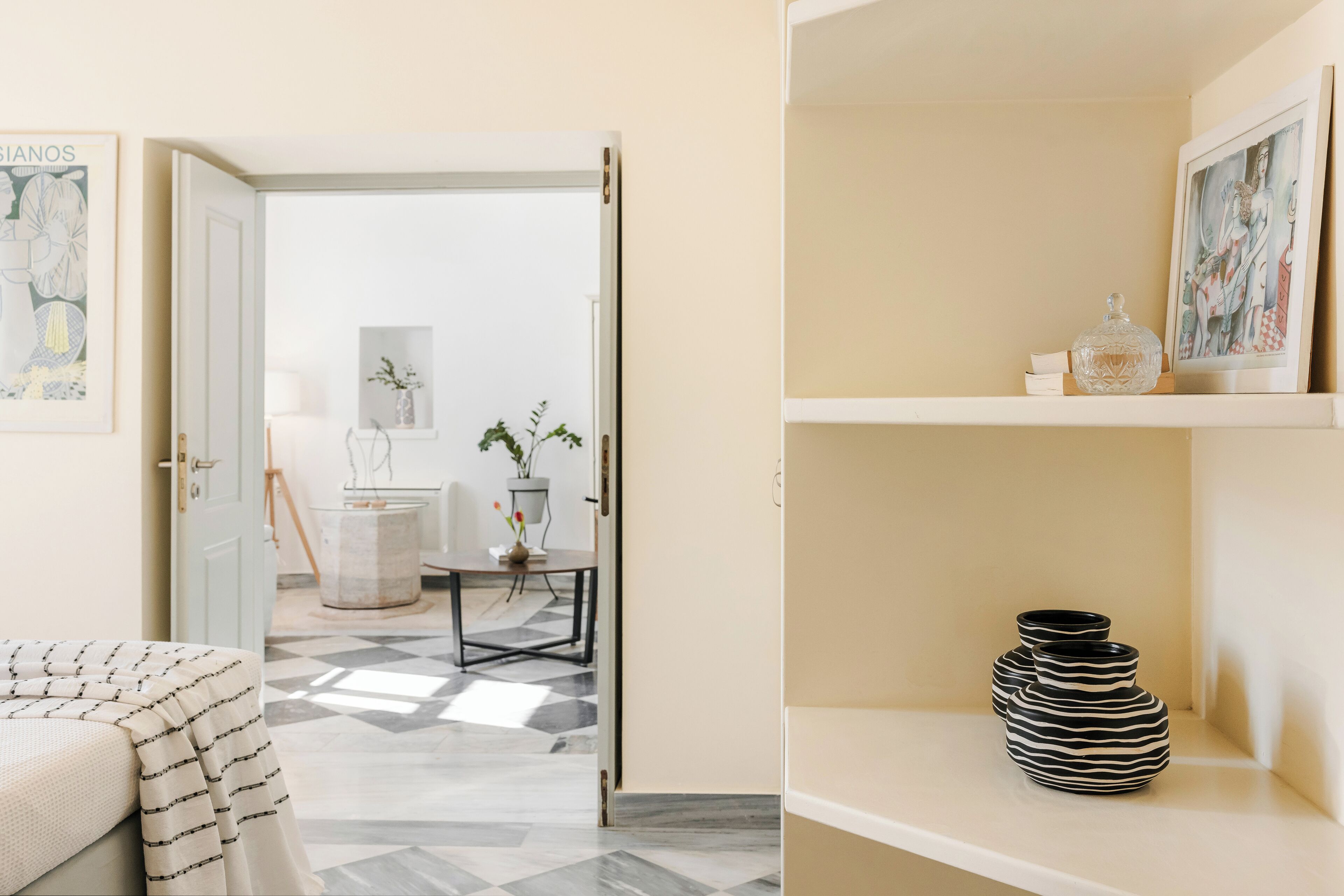 Quarto com cama, porta que dá para outro espaço e prateleira com objetos decorativos.