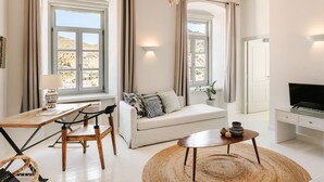 A modern living room with a sofa, a wooden table, a TV, and large windows with curtains. - Hydra Hotel (Hydra)