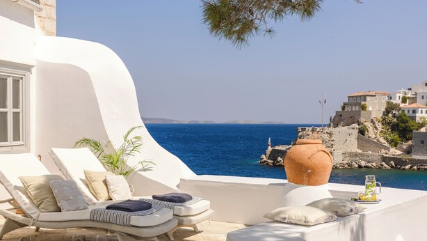A seaside terrace with lounge chairs, a large terracotta pot, and a view of the sea and historic buildings. - Hydrea Exclusive Hospitality (Hydra)