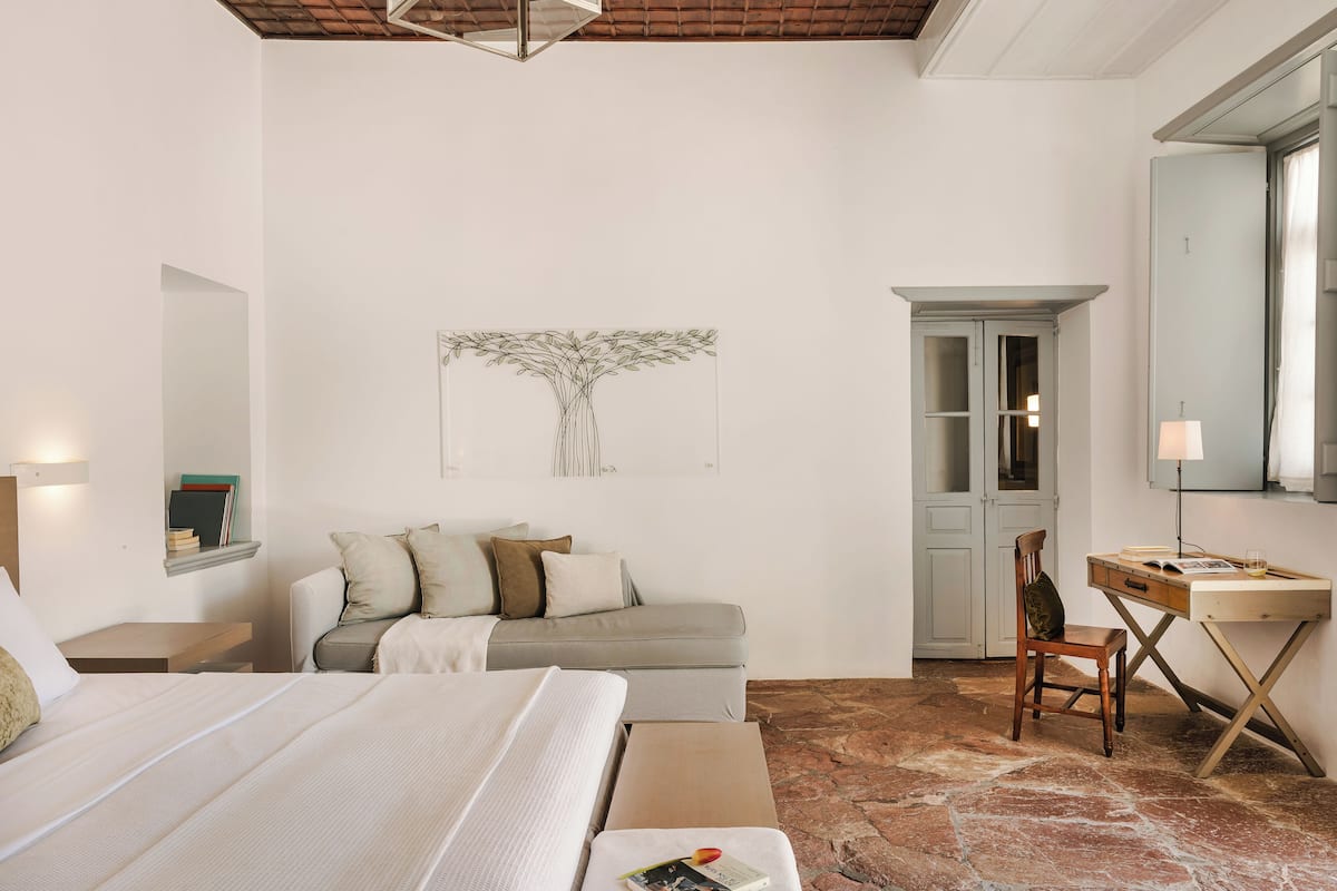 A bedroom with a bed, a sofa, a desk, and a chair. There is a wardrobe and a mirror on the wall.