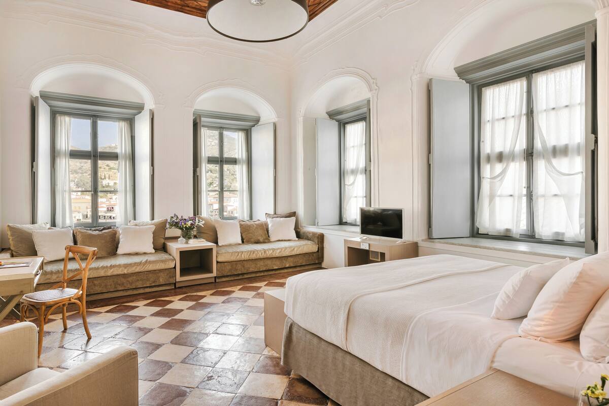 A spacious bedroom with a large bed, a sofa, a TV, and multiple windows with curtains.