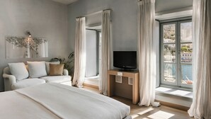 A modern bedroom with a large bed, a TV on a wooden stand, a sofa, and a view of the sea through large windows. - Hydrea Exclusive Hospitality (Hydra)