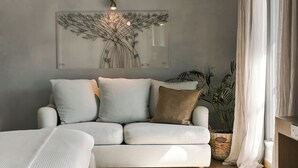 A modern bedroom with a light-colored sofa, a bedside table, and a large window with curtains. - Hydrea Exclusive Hospitality (Hydra)