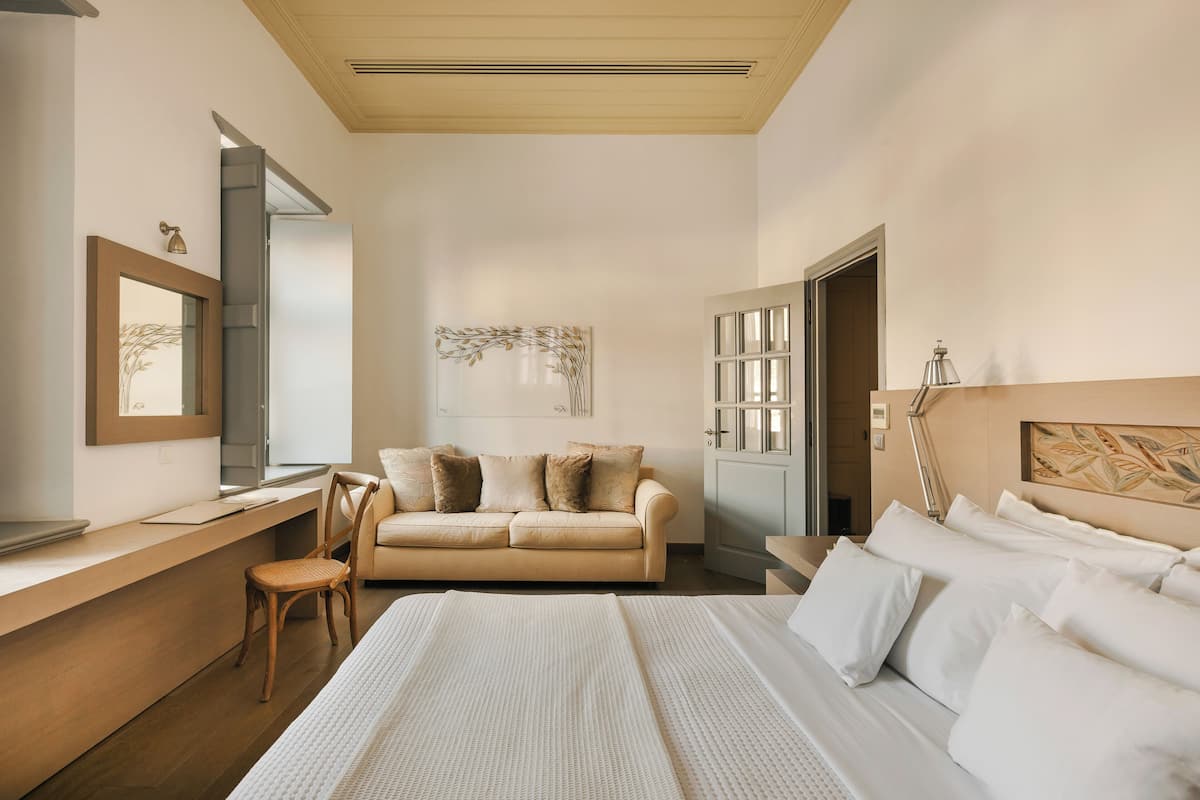 A modern hotel room with a large bed, a sitting area with a sofa and a chair, a desk, and a wardrobe.