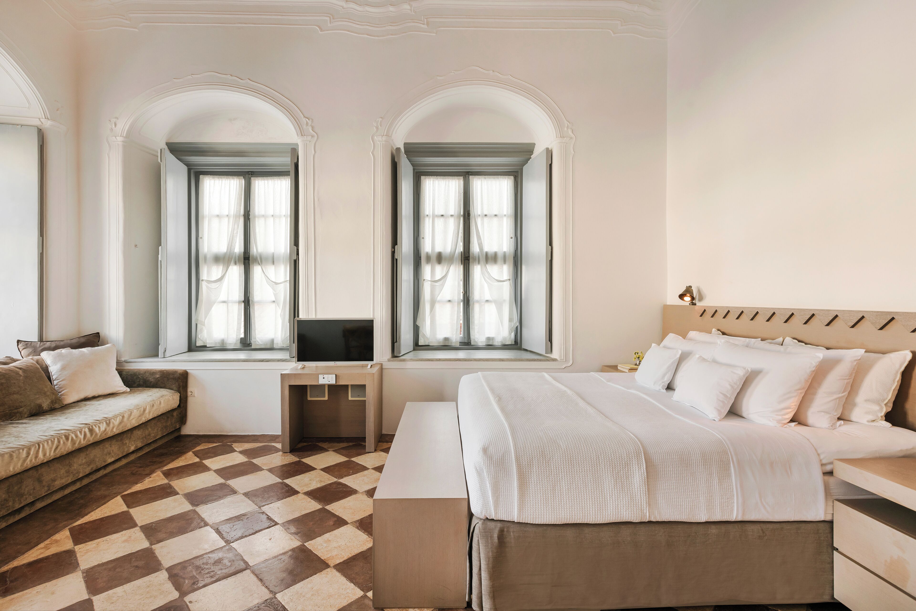 Una camera d'albergo con pavimento a scacchi, un letto con lenzuola bianche, un divano e due finestre con tende.