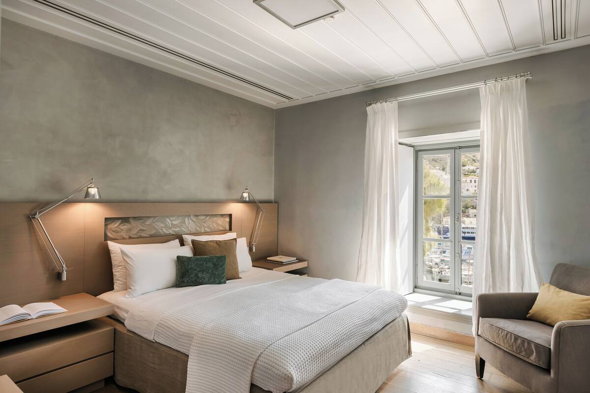 A modern bedroom with a large bed, bedside tables, a nightstand, a reading lamp, and a view of the outside through a window with curtains.