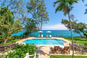 A pool area with lounge chairs and umbrellas, surrounded by lush greenery and overlooking a clear blue sea.