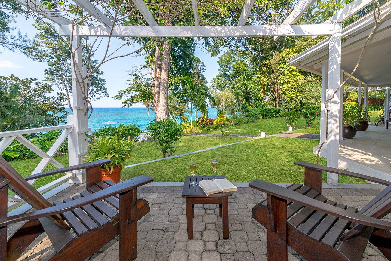 A covered outdoor seating area with wooden chairs and a table, overlooking a lush garden and the ocean.