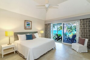 A hotel room with a large bed, two bedside tables with lamps, a ceiling fan, a balcony with a view of the ocean, and a chair.