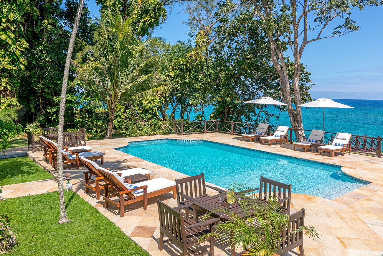 Piscina retangular com deck ladrilhado, rodeada por espreguiçadeiras e mesas de madeira, com vista para o oceano.