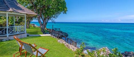 A gazebo with a view of the ocean, a grassy area with two chairs, and a wooden walkway leading to the water.