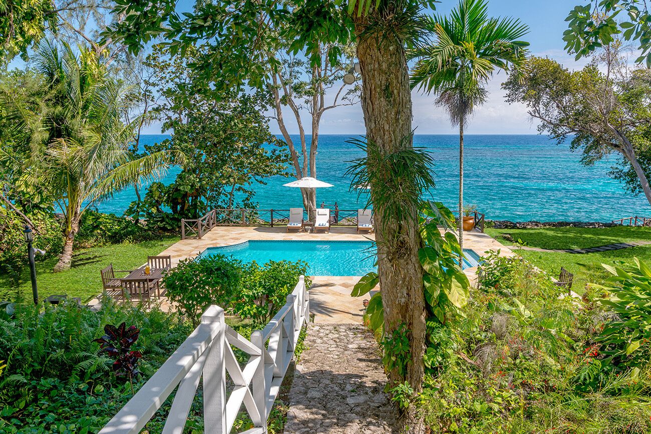 A pool area with a view of the ocean, surrounded by lush greenery and a white fence.