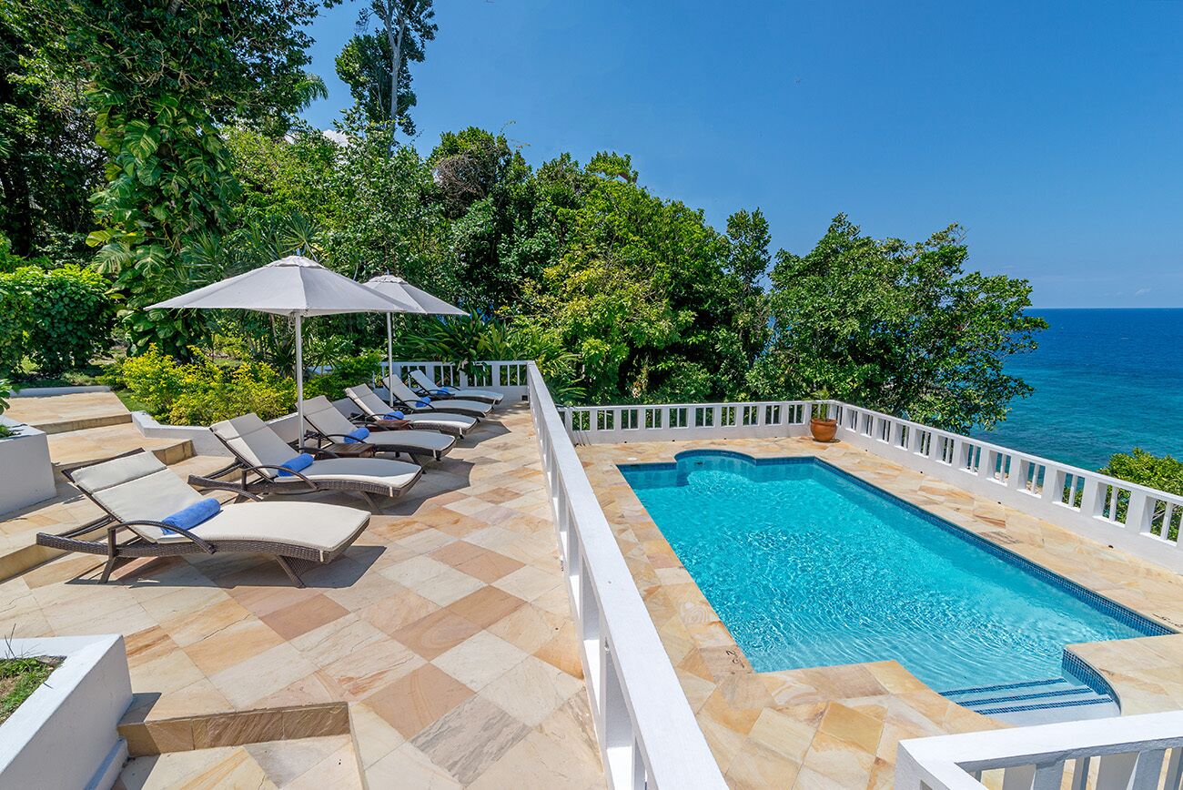 A poolside area with lounge chairs and umbrellas, surrounded by lush greenery and overlooking the sea.