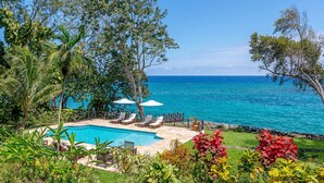 Zona de piscina con sillas de descanso y sombrillas, rodeada de vegetación tropical y vistas a un mar azul y transparente.