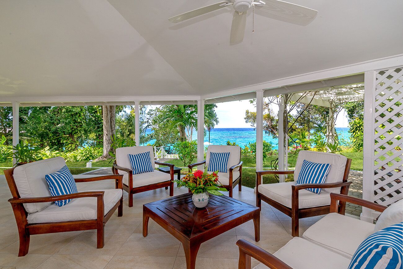 A covered outdoor seating area with wooden furniture and a ceiling fan, overlooking a lush garden and the ocean.