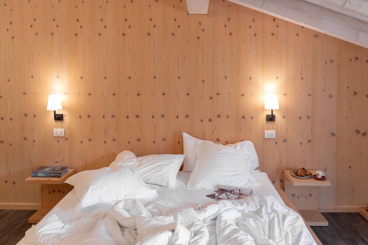 Un dormitorio con pared de madera, una cama con ropa blanca, dos mesitas de noche con lámparas y un estante pequeño con un libro y un vaso.