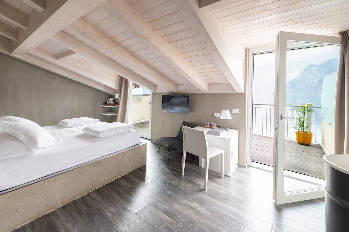 Un dormitorio con cama, escritorio y silla. Hay un televisor en la pared y un balcón con vista a las montañas.