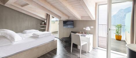 A bedroom with a bed, a desk, and a chair. There is a TV on the wall and a balcony with a view of mountains.