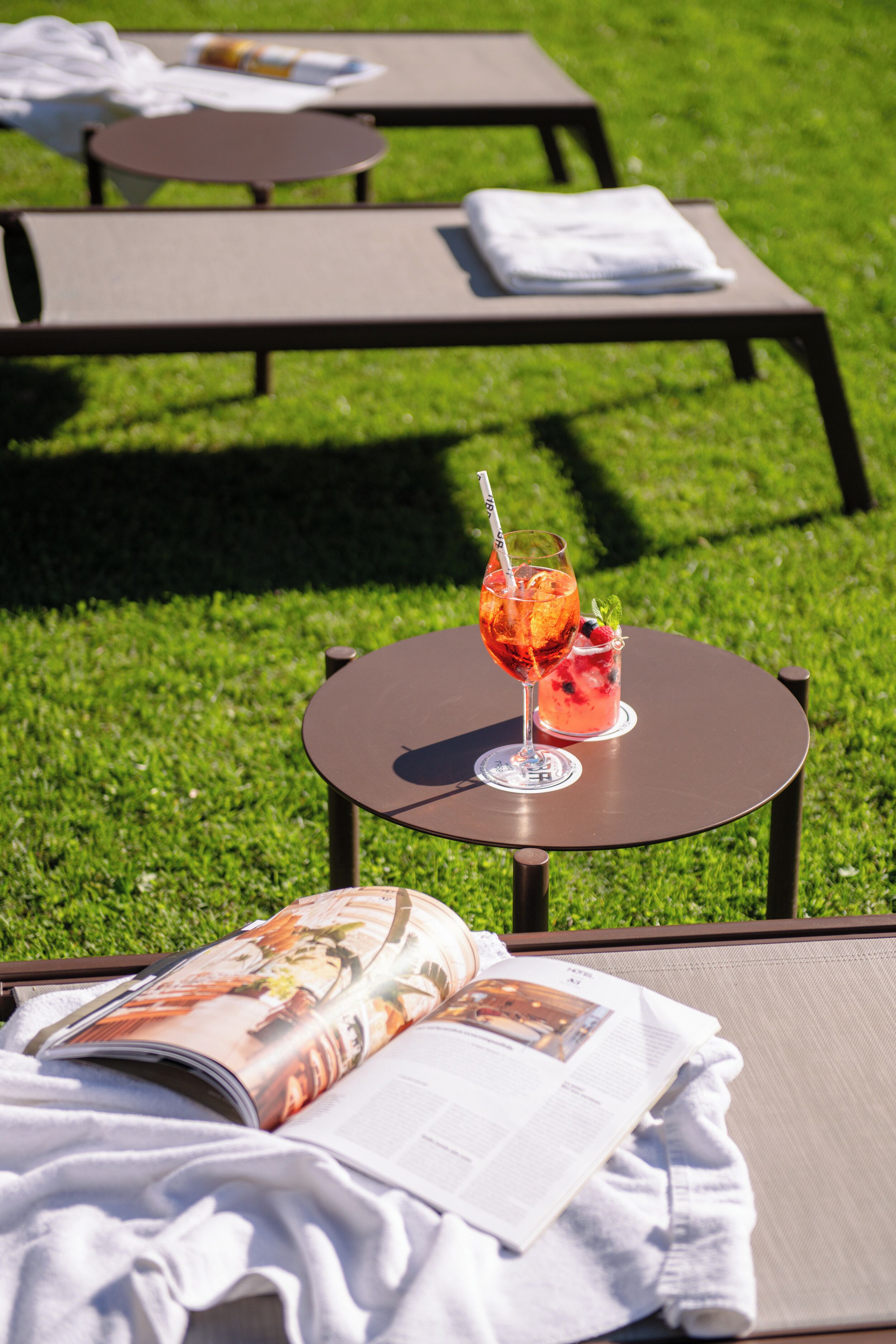 A grassy outdoor area with lounge chairs and a table with a drink and magazine.