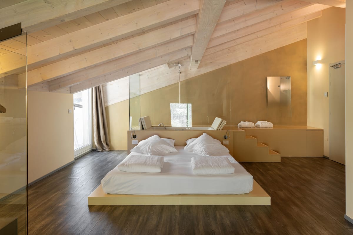 Un dormitorio moderno con cama estilo loft, vigas de madera, un espejo grande y vistas al exterior.
