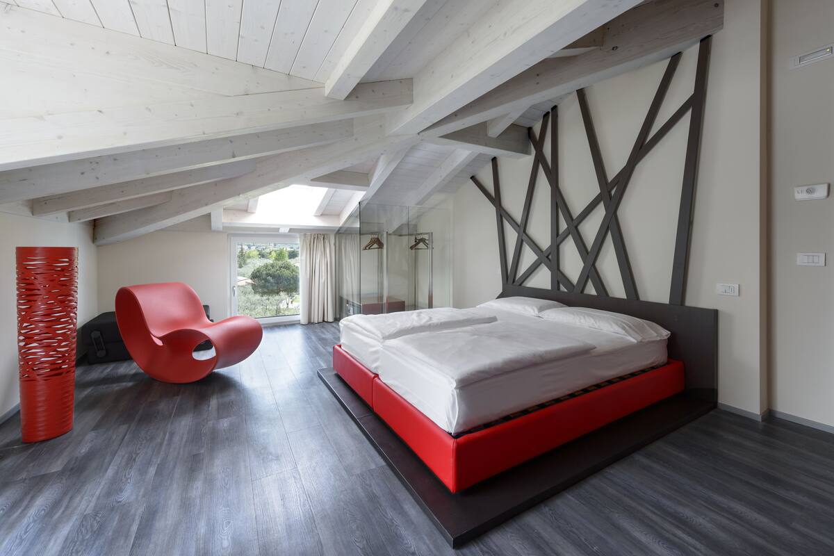 Un dormitorio moderno con techo inclinado, una cama roja y negra, una silla roja singular y un amplio ventanal con vistas a la vegetación.