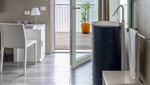 Una habitación moderna con techo de madera, un escritorio blanco con lámpara, un barril negro grande y vistas a montañas a través de una puerta abierta.