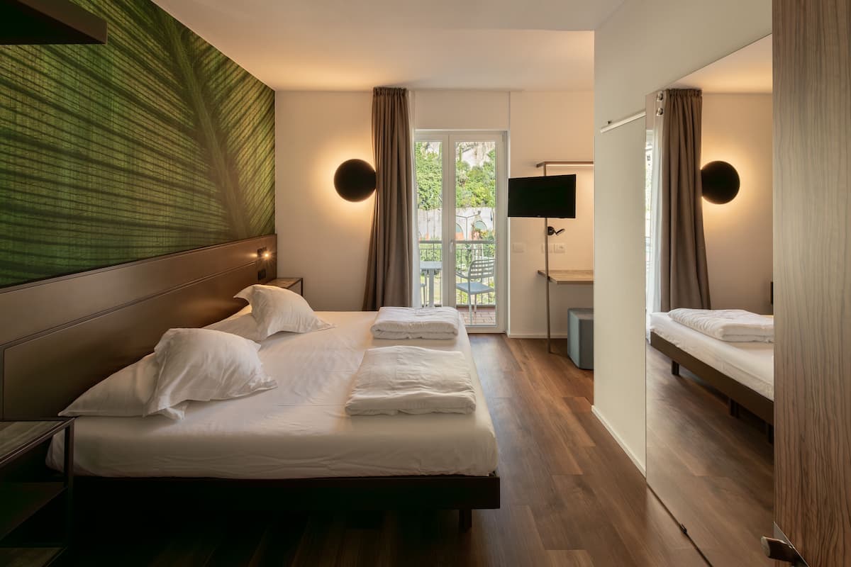 Habitación de hotel con una cama grande, televisión, escritorio y balcón con vistas a zonas verdes.