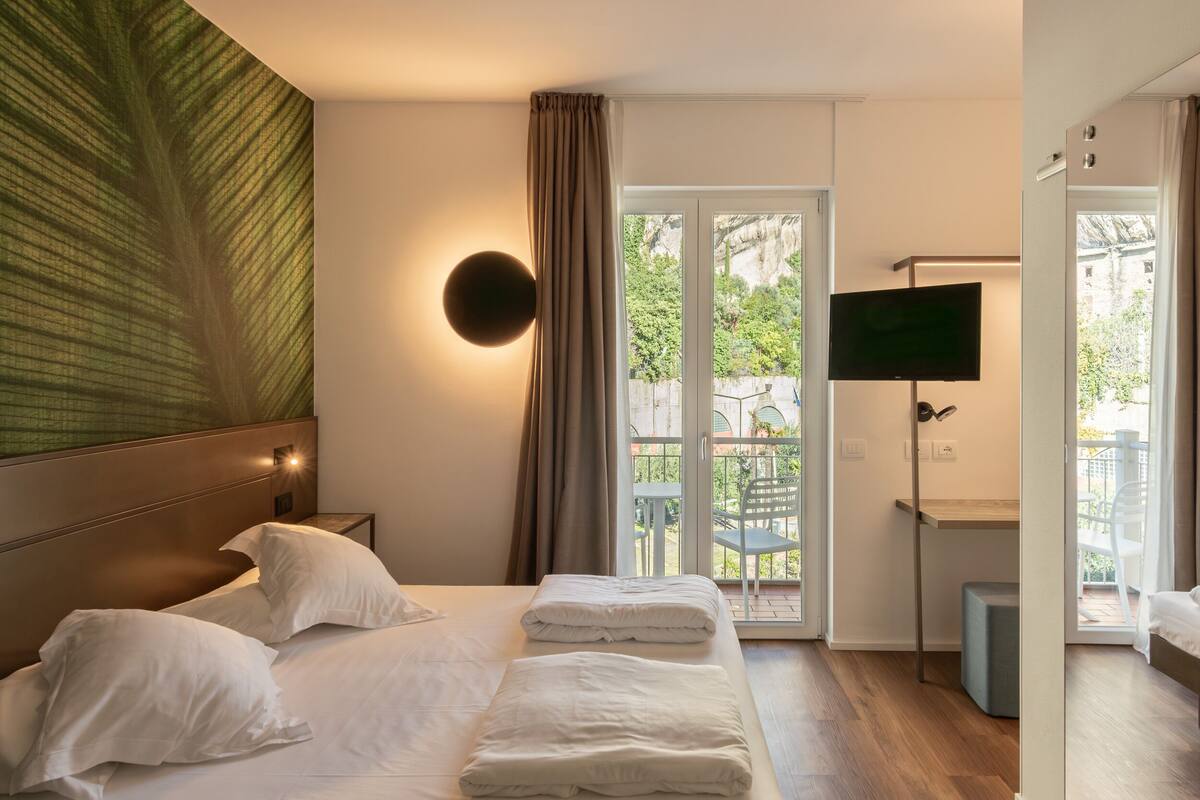 Habitación de hotel moderna con una cama grande, un televisor de pantalla plana, un escritorio y un balcón con vistas a zonas verdes.