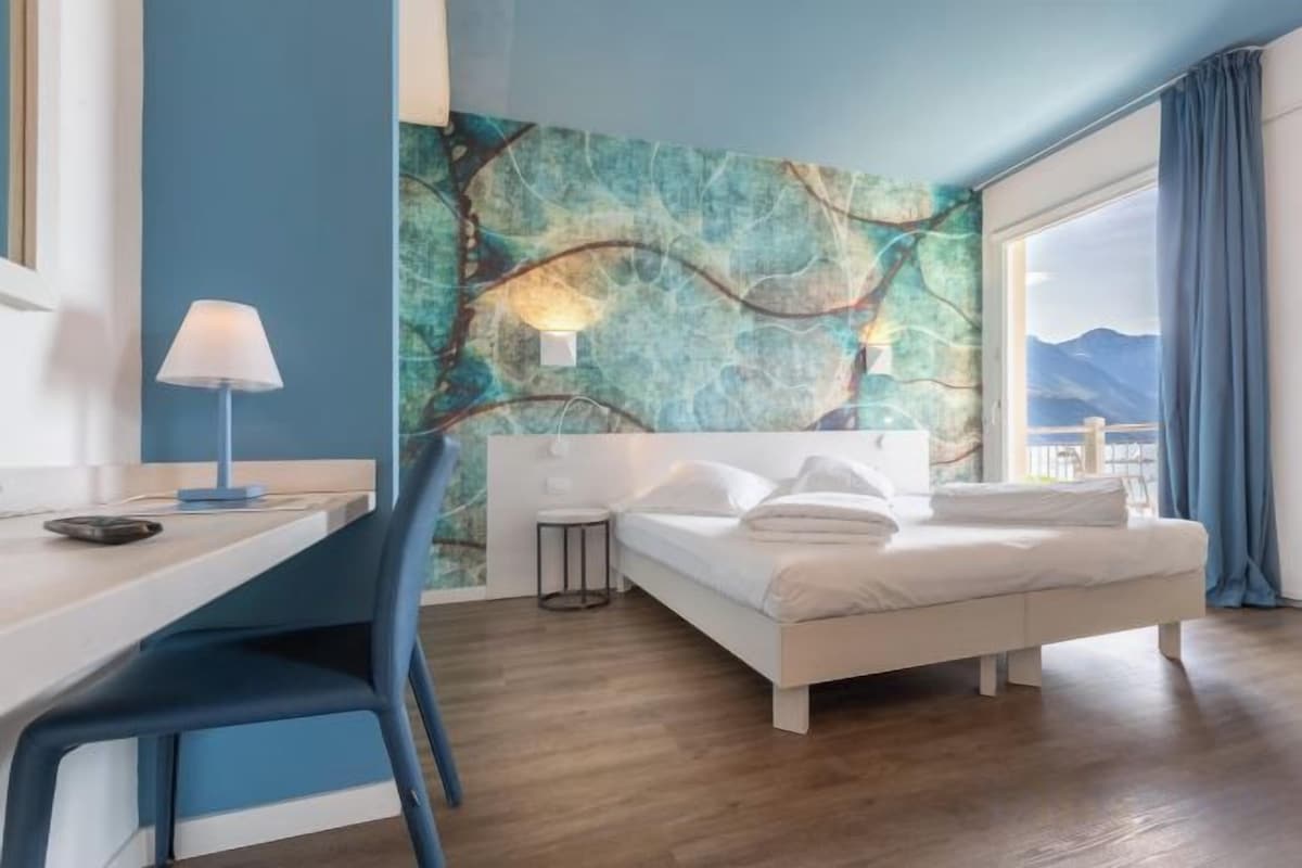 Habitación de hotel con cama, escritorio, silla, lámpara y un ventanal con vistas.