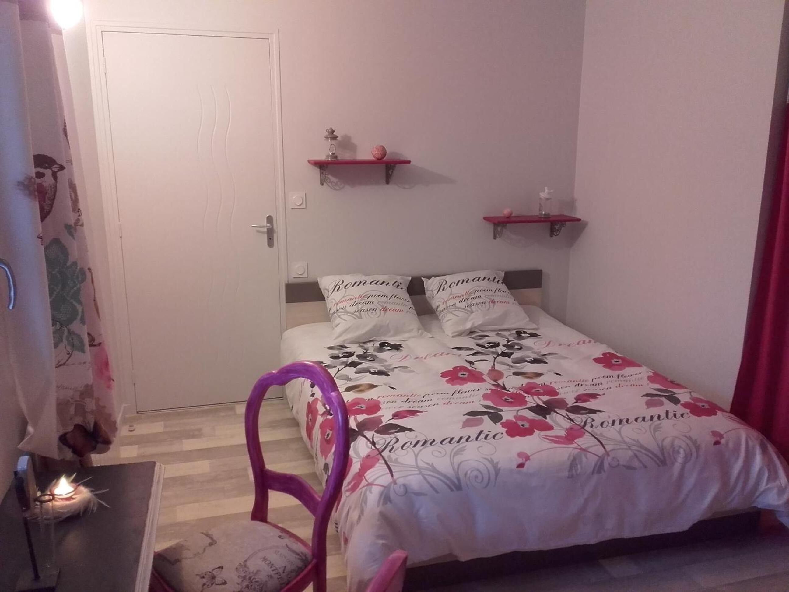 Une chambre à coucher avec un lit, une literie à motifs floraux, deux tables de chevet, une chaise et une porte.