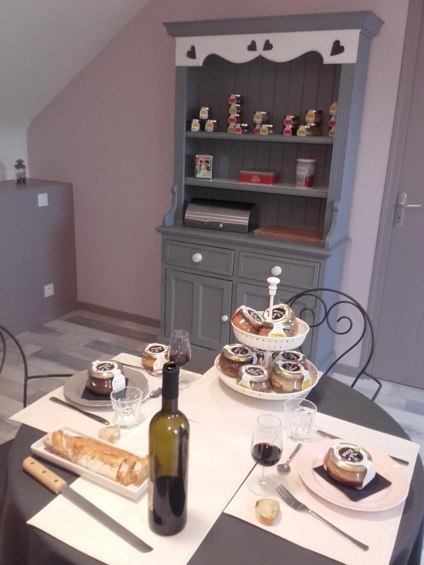 A dining table set with a bottle of wine, glasses, plates with food, and a decorative shelf with jars and a small figurine.