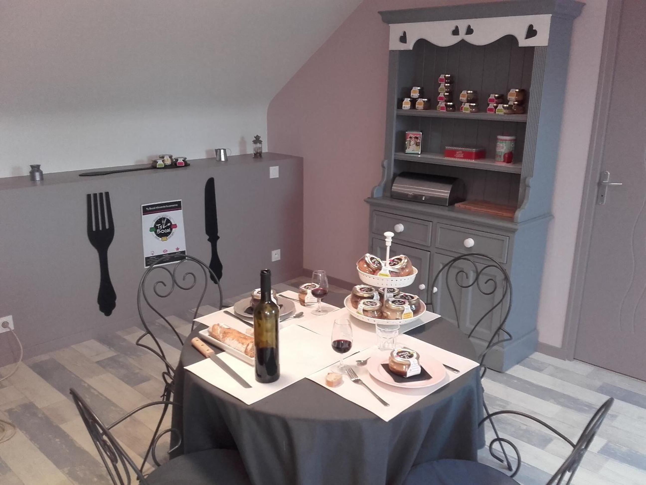 Un comedor con una mesa dispuesta para dos personas, que incluye una botella de vino, hogaza de pan y una bandeja de postres.
