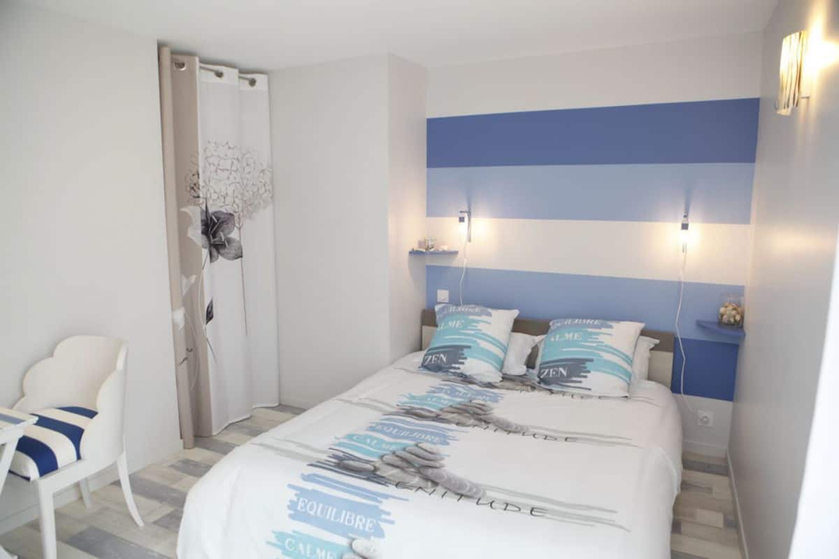 A bedroom with a bed, a chair, a nightstand, and a wall-mounted light fixture.