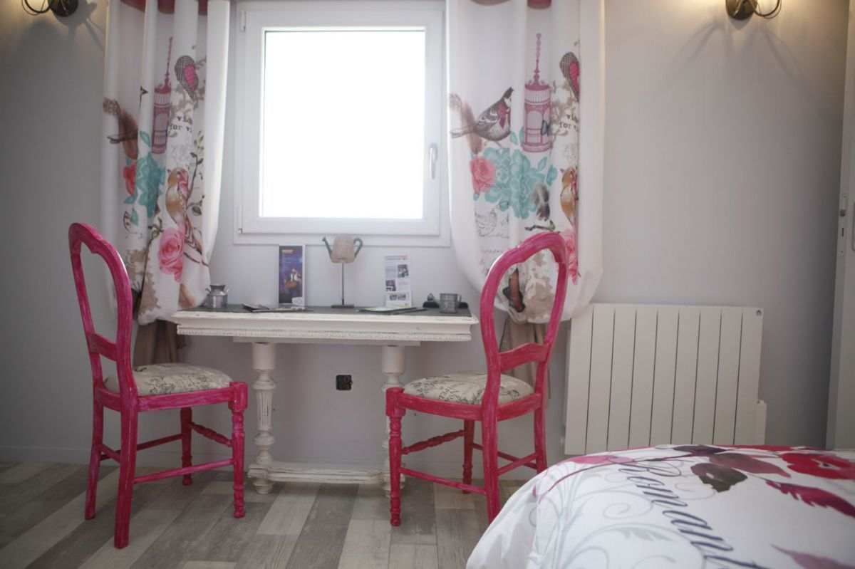 A bedroom with a white bed, a small desk with a chair, a radiator, and floral curtains.
