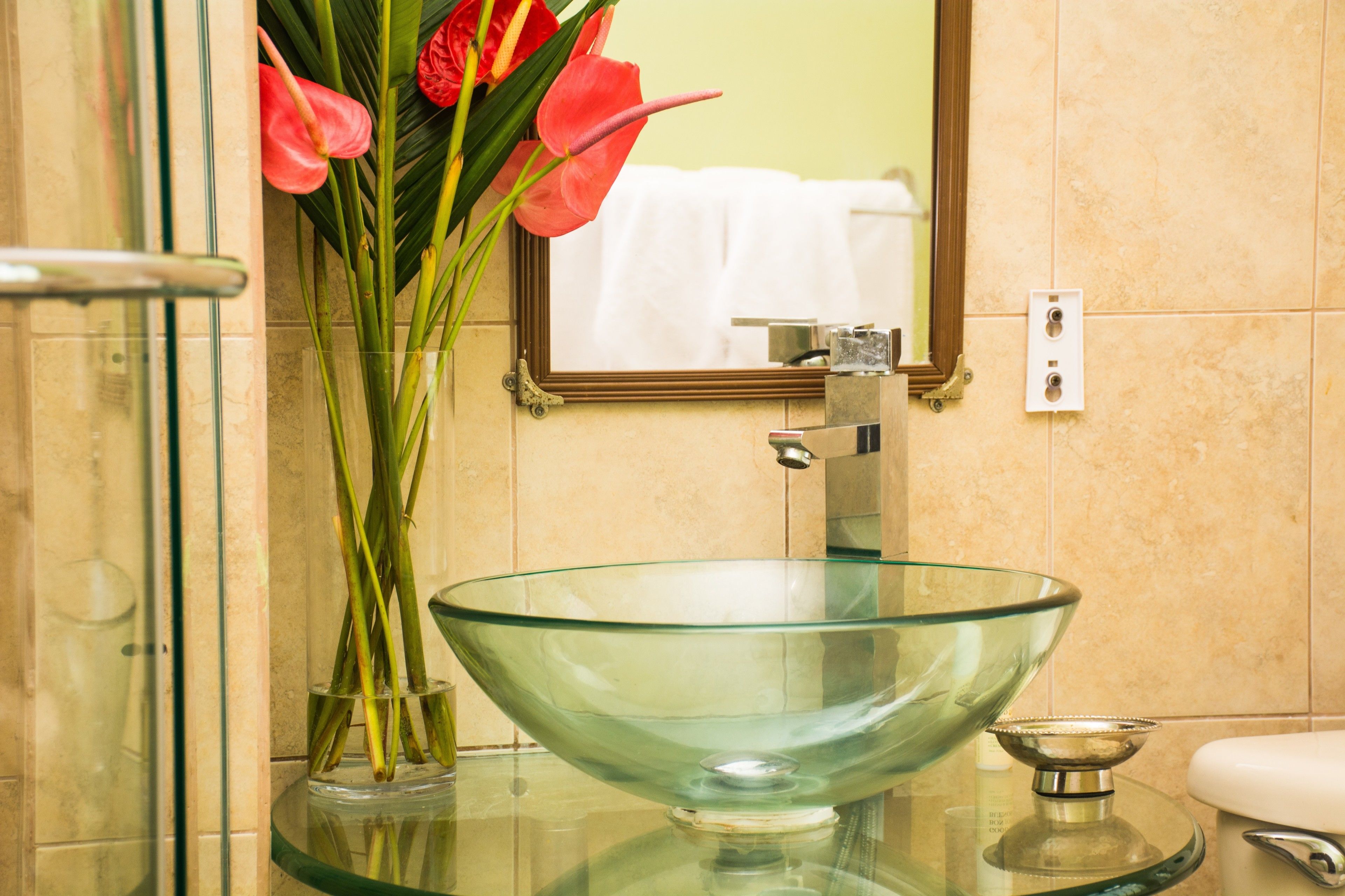 Une salle de bain avec un lavabo en forme de bol en verre, un miroir et un vase rempli de fleurs rouges.