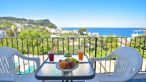 Un balcon avec une table dressée pour deux personnes, une assiette de fruits et deux verres de jus, donnant sur une ville côtière et la mer.