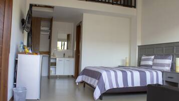 A bedroom with a bed, a desk, a mirror, and a kitchenette.