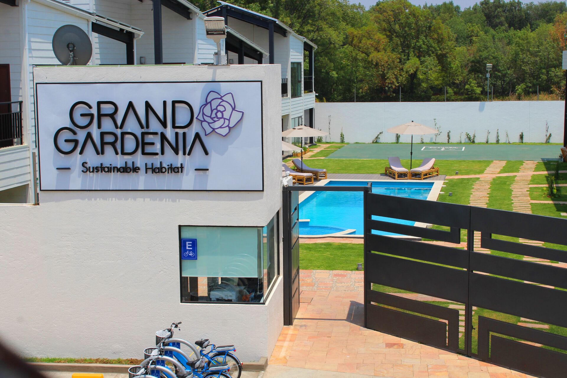 A sign for Grand Gardenia Sustainable Habitat with a view of a swimming pool and outdoor seating.