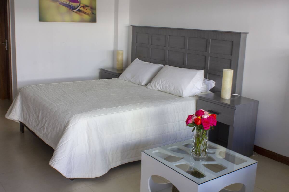 Een netjes opgemaakt slaapkamer met een bed, nachtkastjes en een kleintje tafeltje met een bloemenvaas.