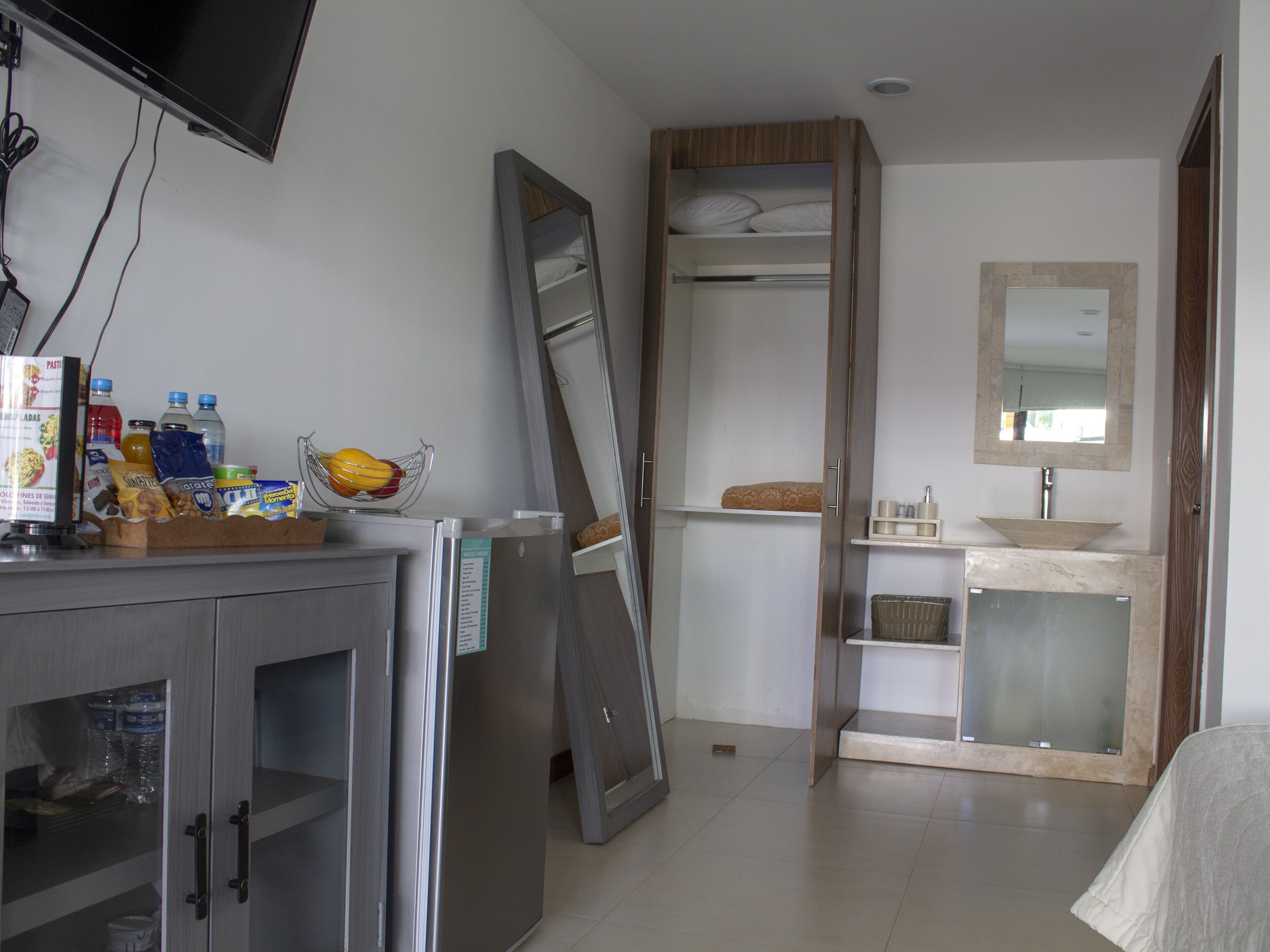 Ein Hotelzimmer mit Bett, Nachttisch mit Fernseher, Kleiderschrank, Badezimmer mit Waschbecken und Spiegel sowie einer kleinen Sitzecke mit einem Sessel.