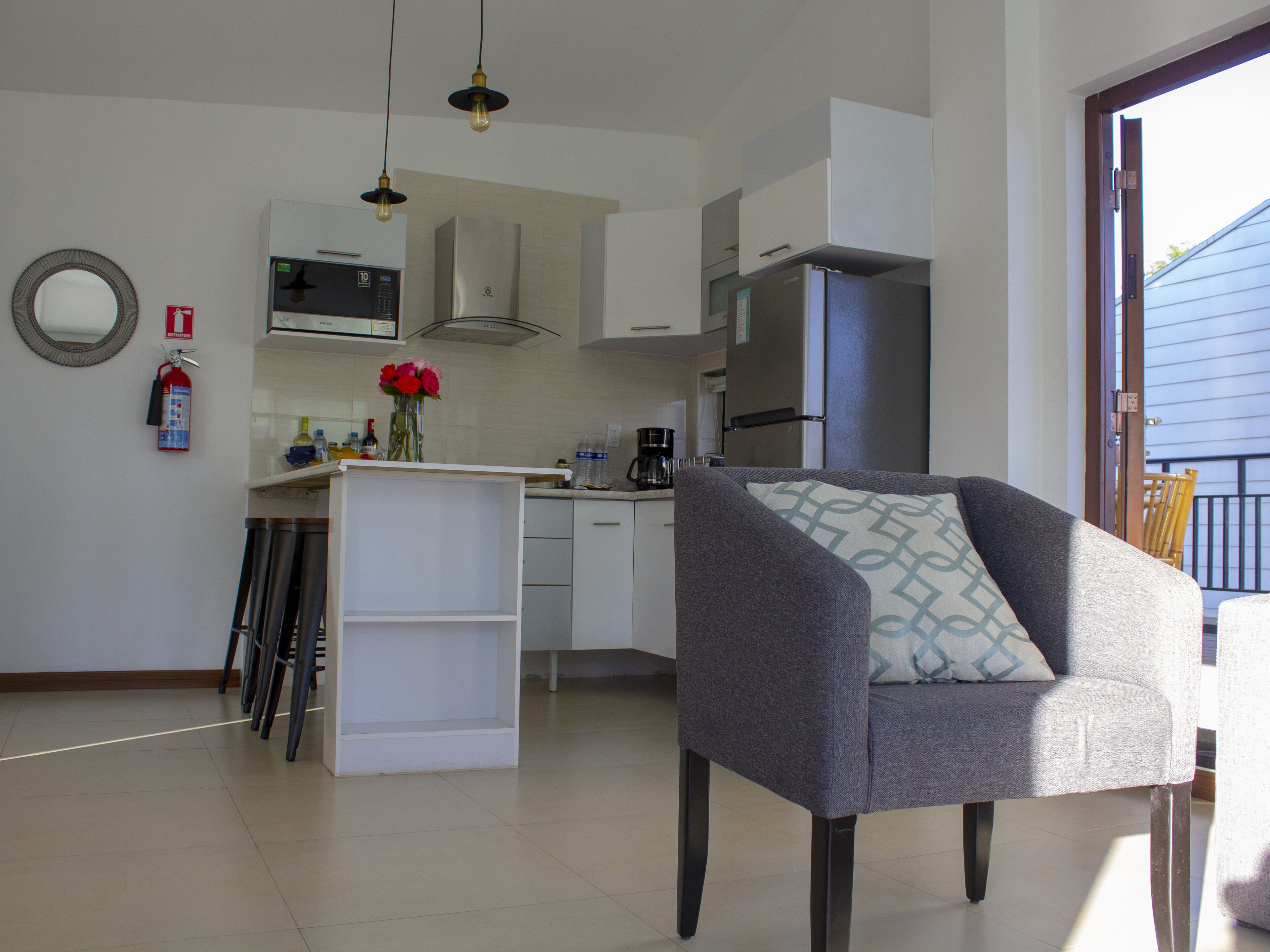 A modern kitchen with white cabinets, a stainless steel refrigerator, and a dining area with a grey armchair and a patterned cushion.