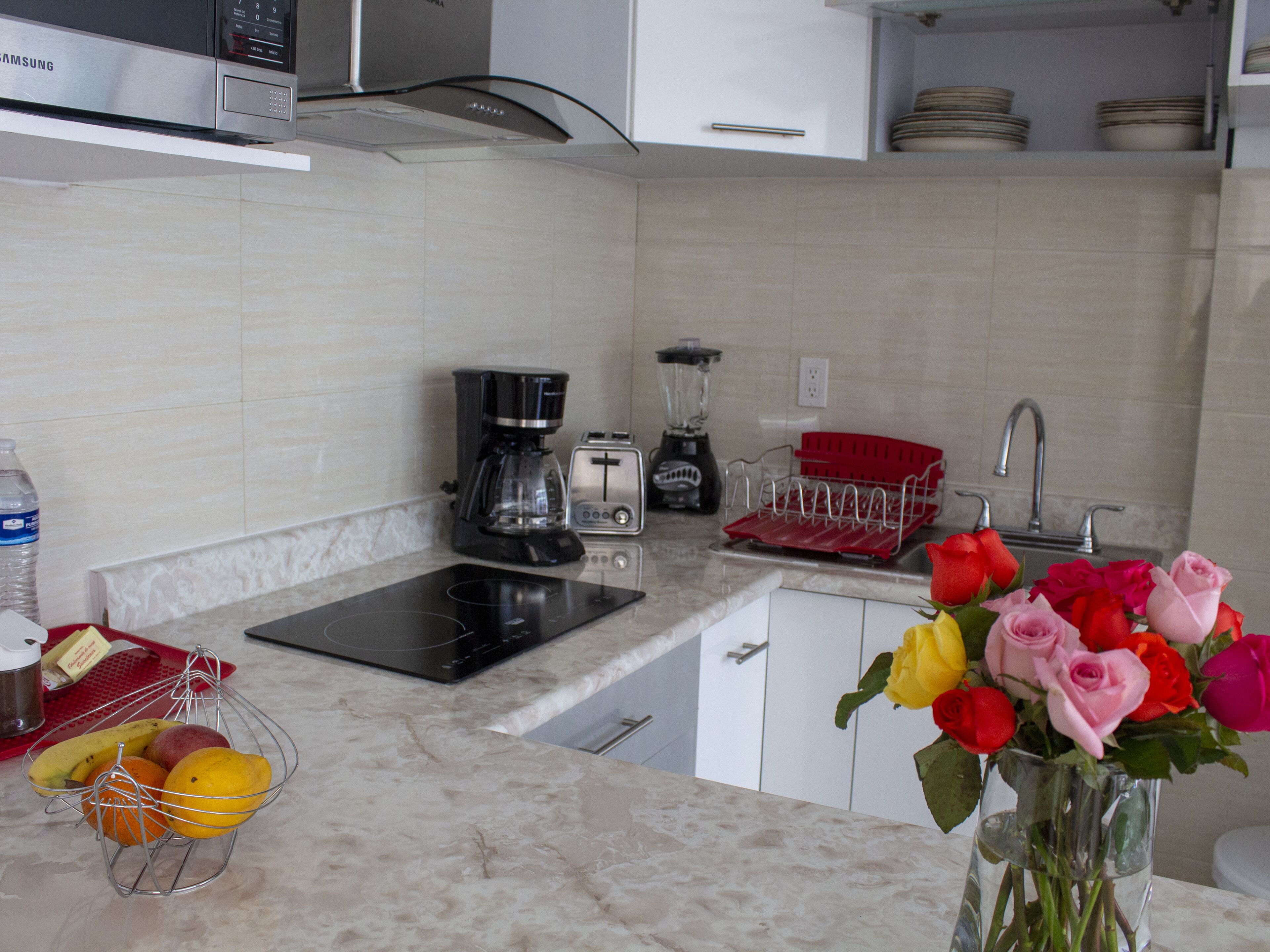 A modern kitchen with a microwave, coffee maker, toaster, and blender. There is a sink with a red dish rack and a vase of flowers on the counter.