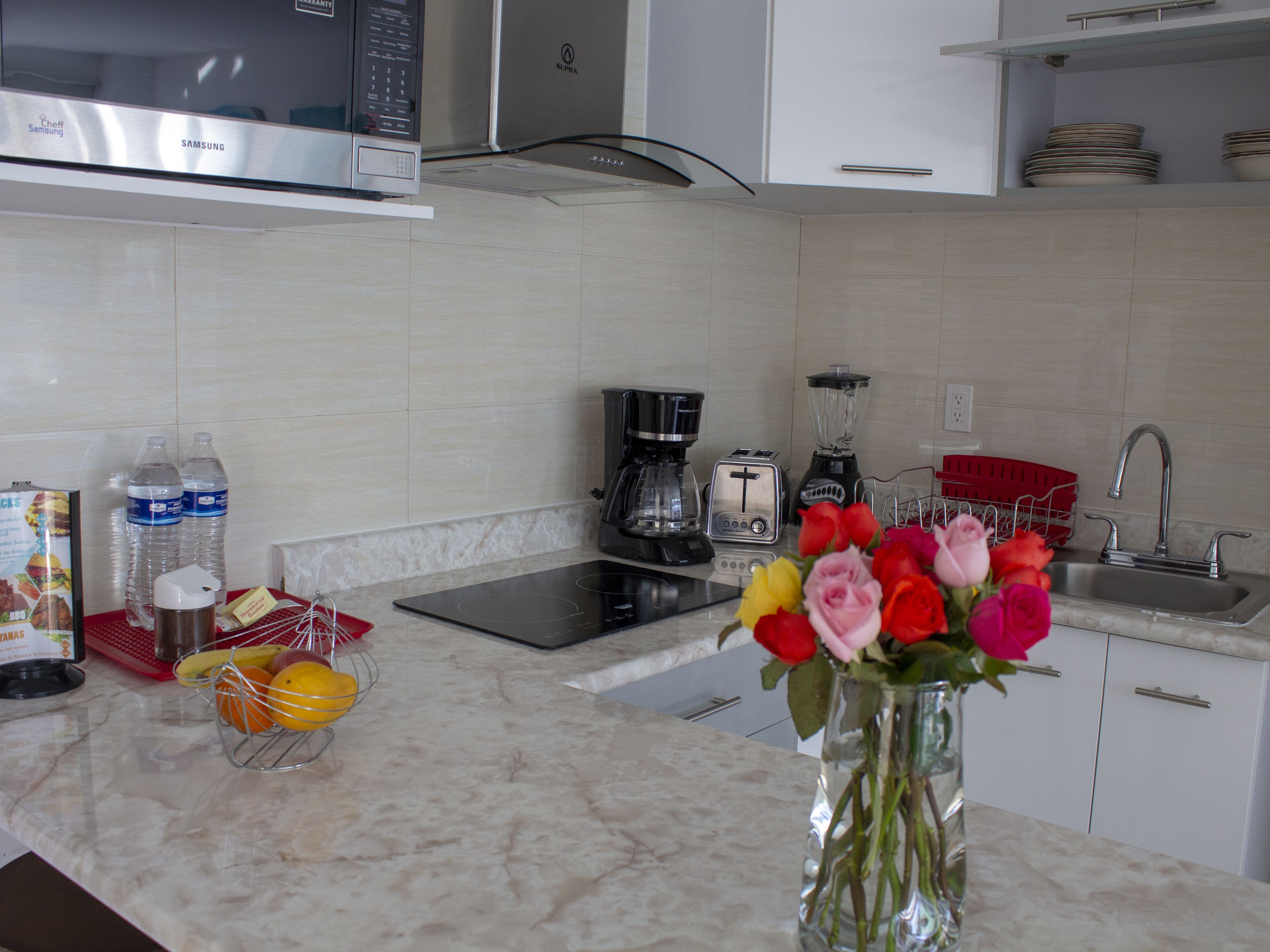 Una cocina moderna con microondas, tostadora, cafetera y lavamanos. Hay botellas de agua, una cesta de frutas y un florero con flores en la encimera.