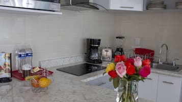 Una cocina moderna con microondas, tostadora, cafetera y lavamanos. Hay botellas de agua, una cesta de frutas y un florero con flores en la encimera.
