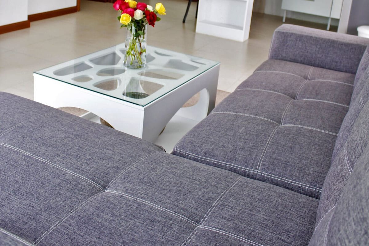 Een moderne woonkamer met een grijze bank, een glazen salontafel met een bloevas, een witte keuken op de achtergrond, en een houten deur.