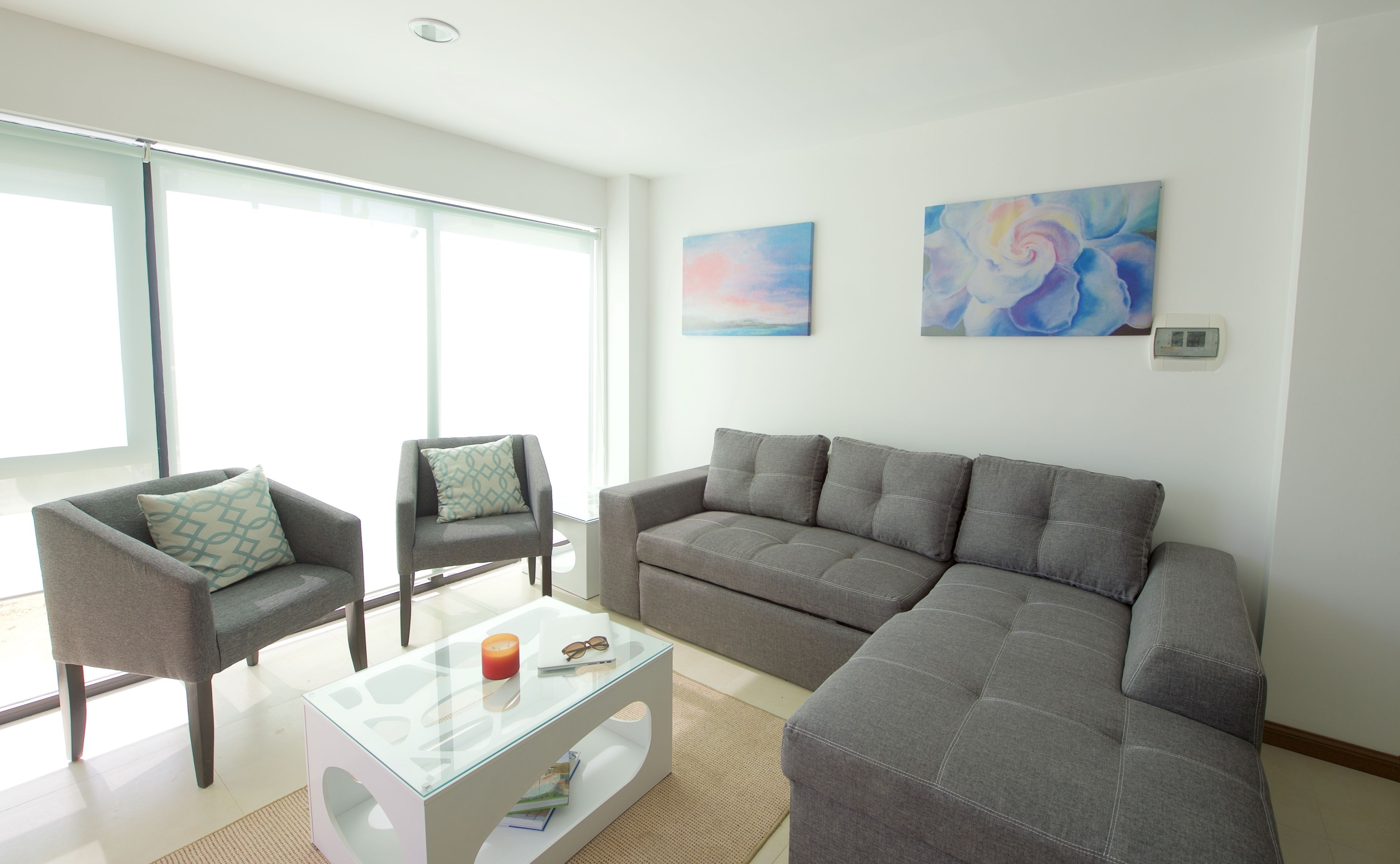 Ein Wohnzimmer mit grauem Sofa, zwei Sesseln, einem Glastisch und zwei abstrakten Bildern an der Wand.