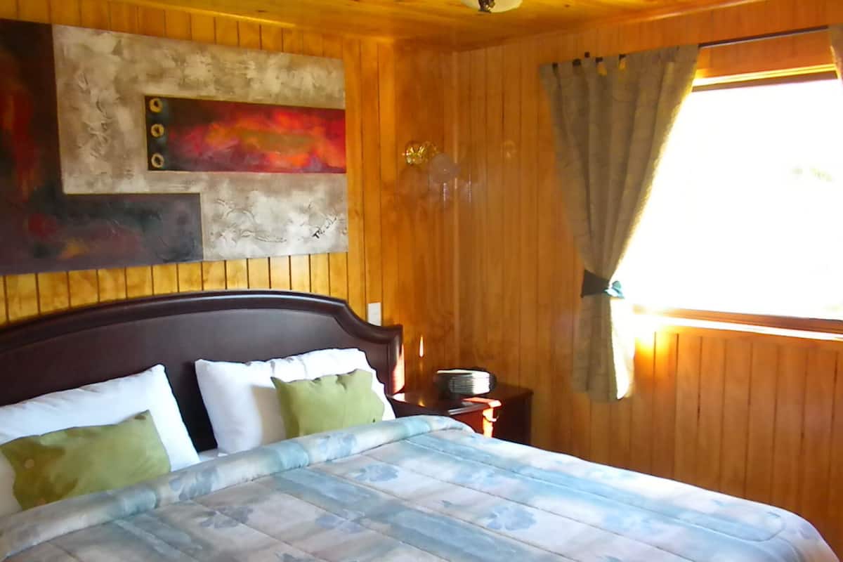 A wooden room with a bed, a painting on the wall, and a window with curtains.